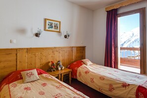 Une chambre avec deux lits, une tête de lit en bois, une petite table sur laquelle sont posés un ours en peluche et une lampe, un tableau encadré au mur et une fenêtre donnant sur des montagnes enneigées.