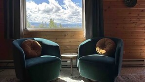 A room with two teal armchairs, a small round table, and a large window with a view of the sky and trees. - Entire Cabin: Step Back in Time at The Piney Lodge (Grand Lake)