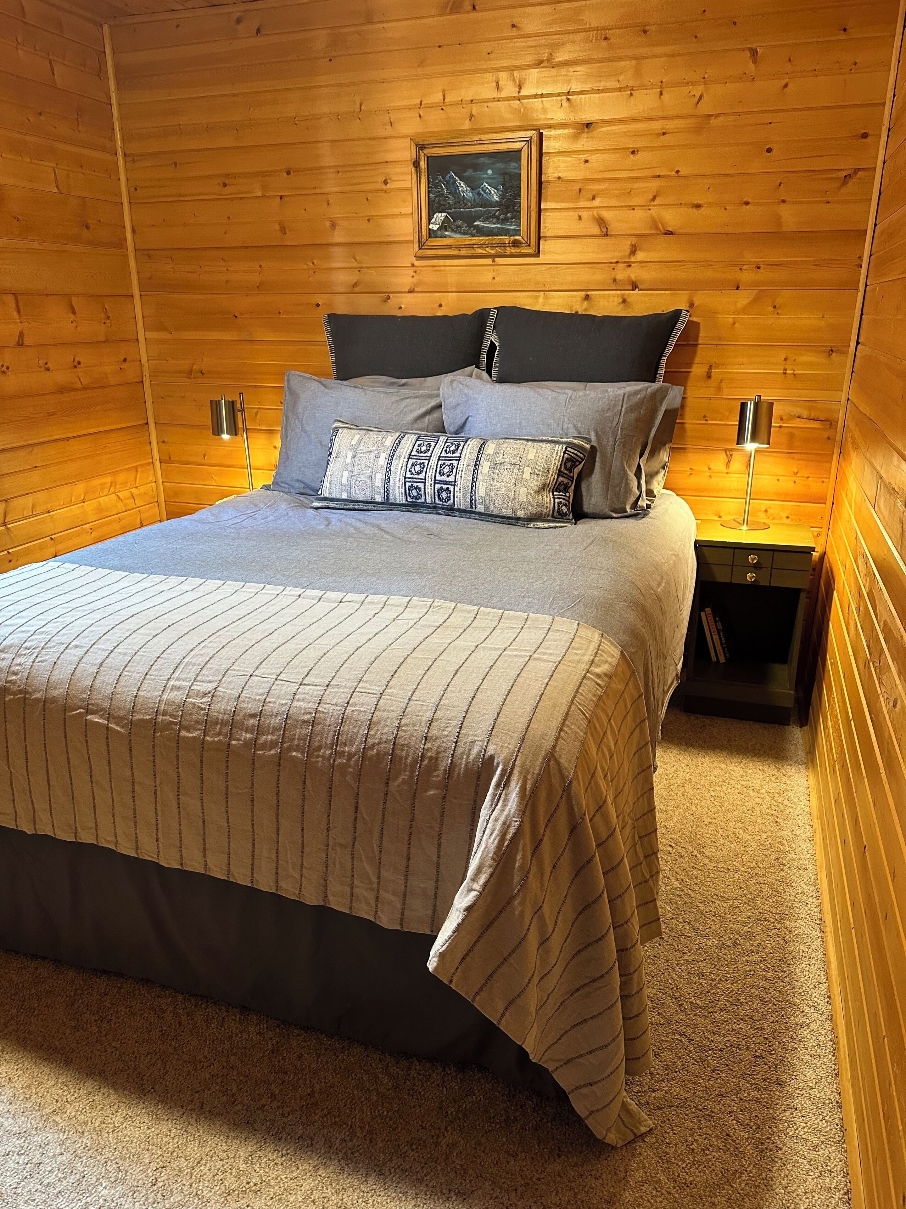 Een knus houten vertrek met een bed, kussens, een nachtkastje, een lamp en een ingelijste foto aan de muur.