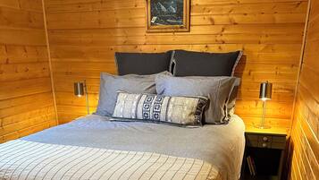 Een knus houten vertrek met een bed, kussens, een nachtkastje, een lamp en een ingelijste foto aan de muur.