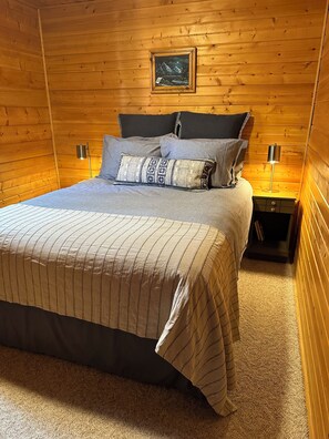 A cozy wooden room with a bed, pillows, a nightstand, a lamp, and a framed picture on the wall. - Entire Cabin: Step Back in Time at The Piney Lodge (Grand Lake)