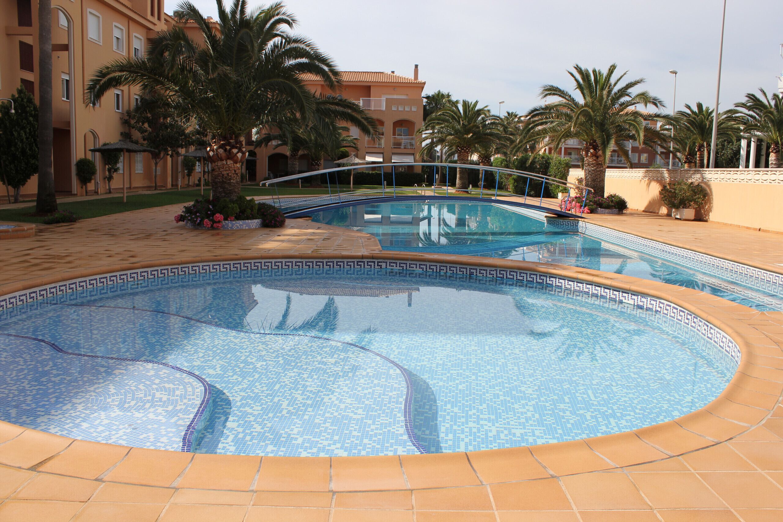 A swimming pool with a tiled edge, surrounded by palm trees and a building with balconies.