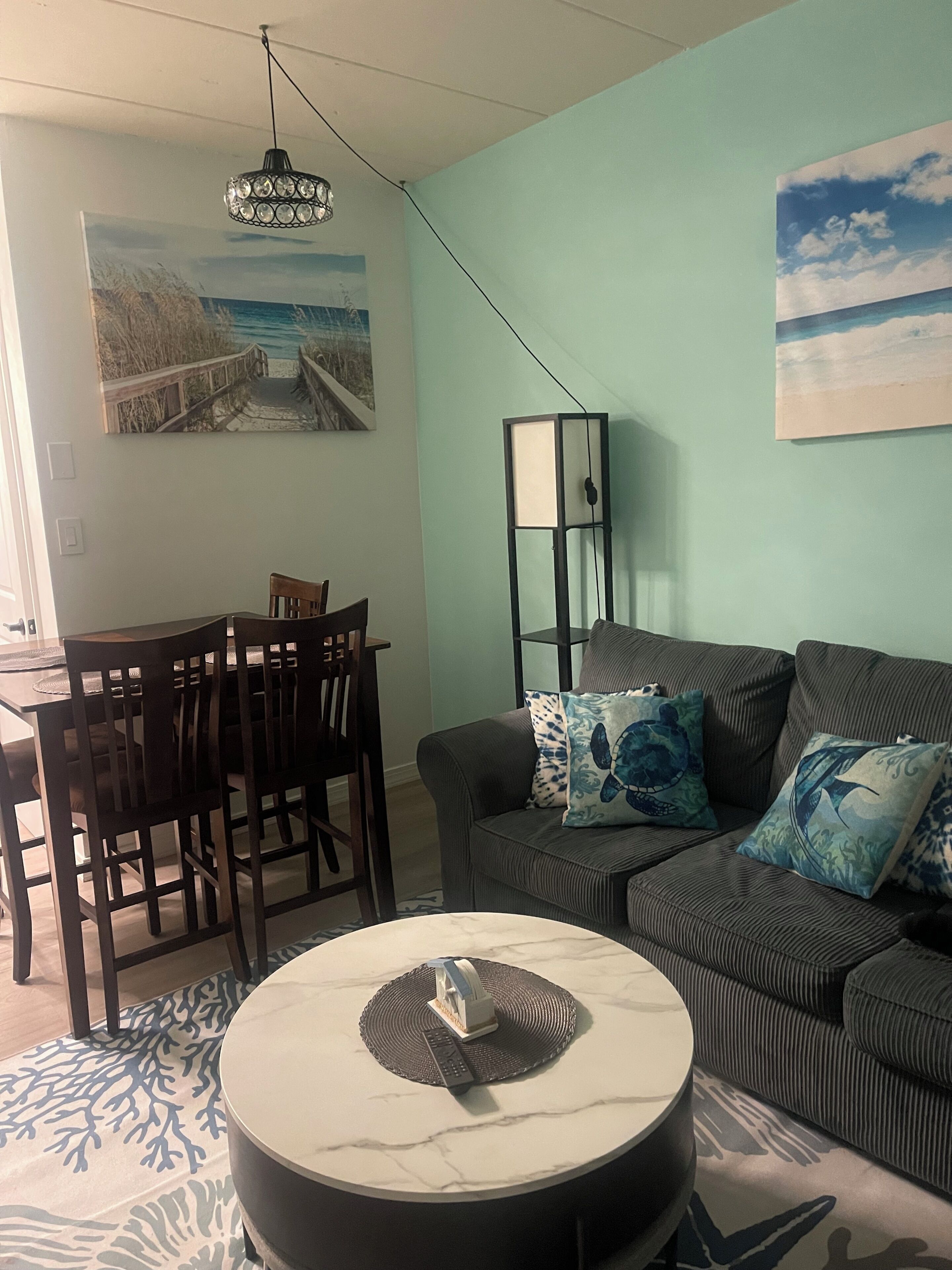 A living room with a sofa, a coffee table, a dining table with chairs, a floor lamp, and wall art.