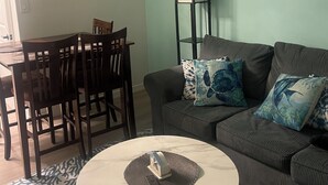 A living room with a sofa, a coffee table, a dining table with chairs, a floor lamp, and wall art. - Excellent location! Close to Famous Boardwalk. One block from the boardwalk. (Wildwood)