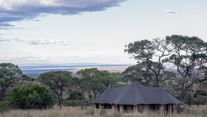 Una cabaña de safari con techo cónico, rodeada de hierba alta y árboles, con vistas a un paisaje lejano.
