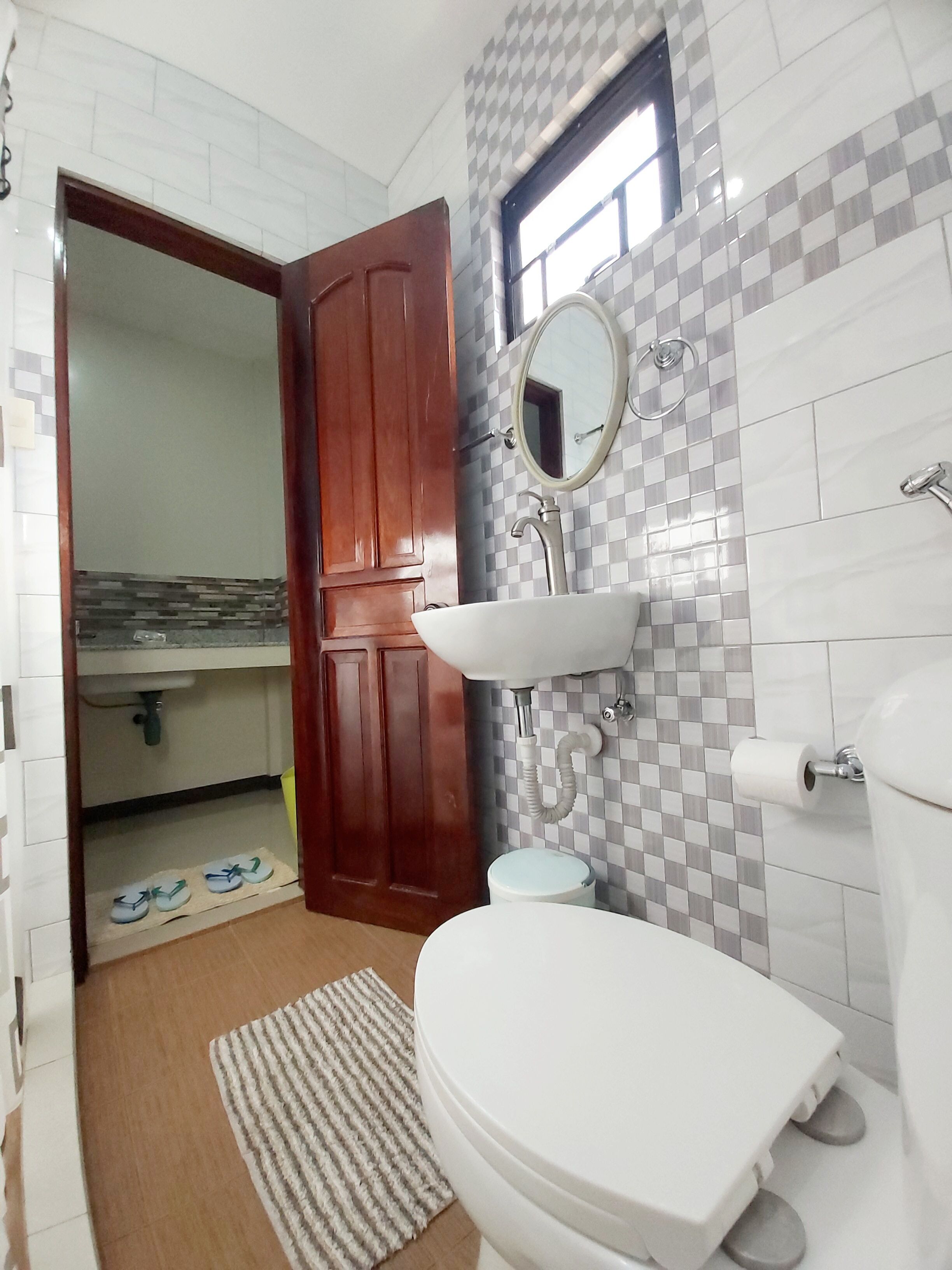 Un baño con inodoro, lavamanos y espejo. Una puerta de madera da a otra habitación.