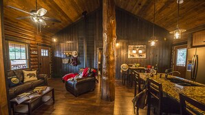 A rustic cabin interior with a wooden floor, leather furniture, a dining area, and a kitchen with a refrigerator.