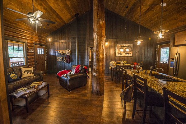 A rustic cabin interior with a wooden floor, leather furniture, a dining area, and a kitchen with a refrigerator.