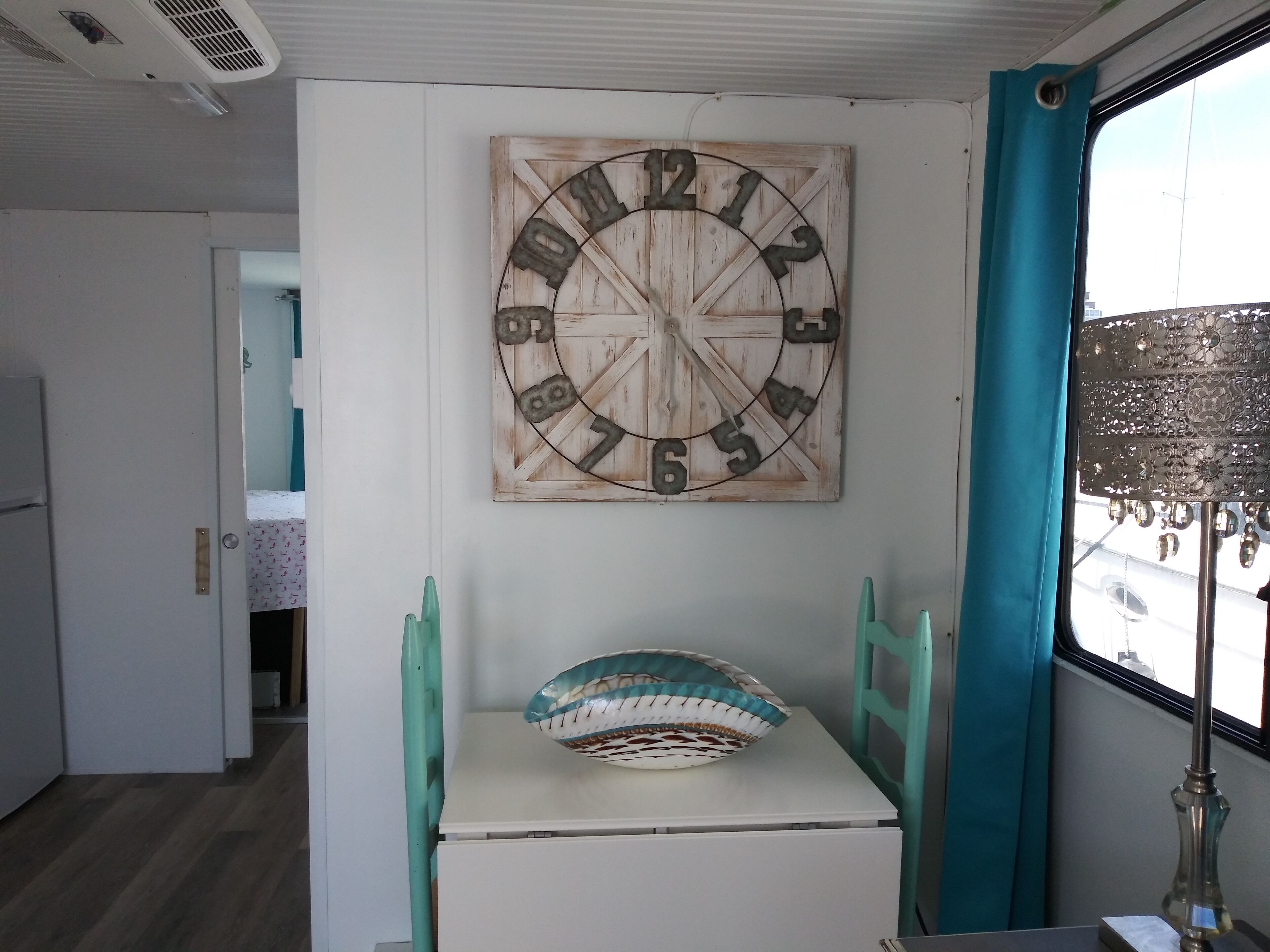 A room with a rustic wooden wall clock, a small table with a patterned cushion, teal chairs, and a window with a view of the outside.