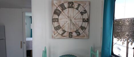 A room with a rustic wooden wall clock, a small table with a patterned cushion, teal chairs, and a window with a view of the outside.