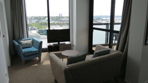 A modern living room with a sofa, a chair, a small table, and a TV. Large windows offer a view of the cityscape. - Ramada Suites By Wyndham Victoria Street West (Auckland)