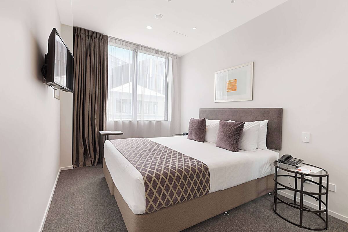 Habitación de hotel con cama, televisión montada en la pared, mesita de noche con teléfono y ventana con cortinas.