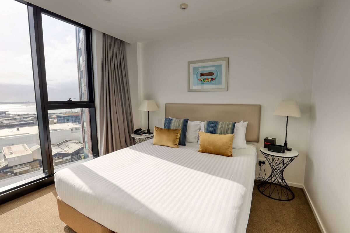 Habitación de hotel con una cama grande, dos mesitas de noche, una mesita auxiliar, un cuadro en la pared y vistas a la ciudad a través de un ventanal amplio.