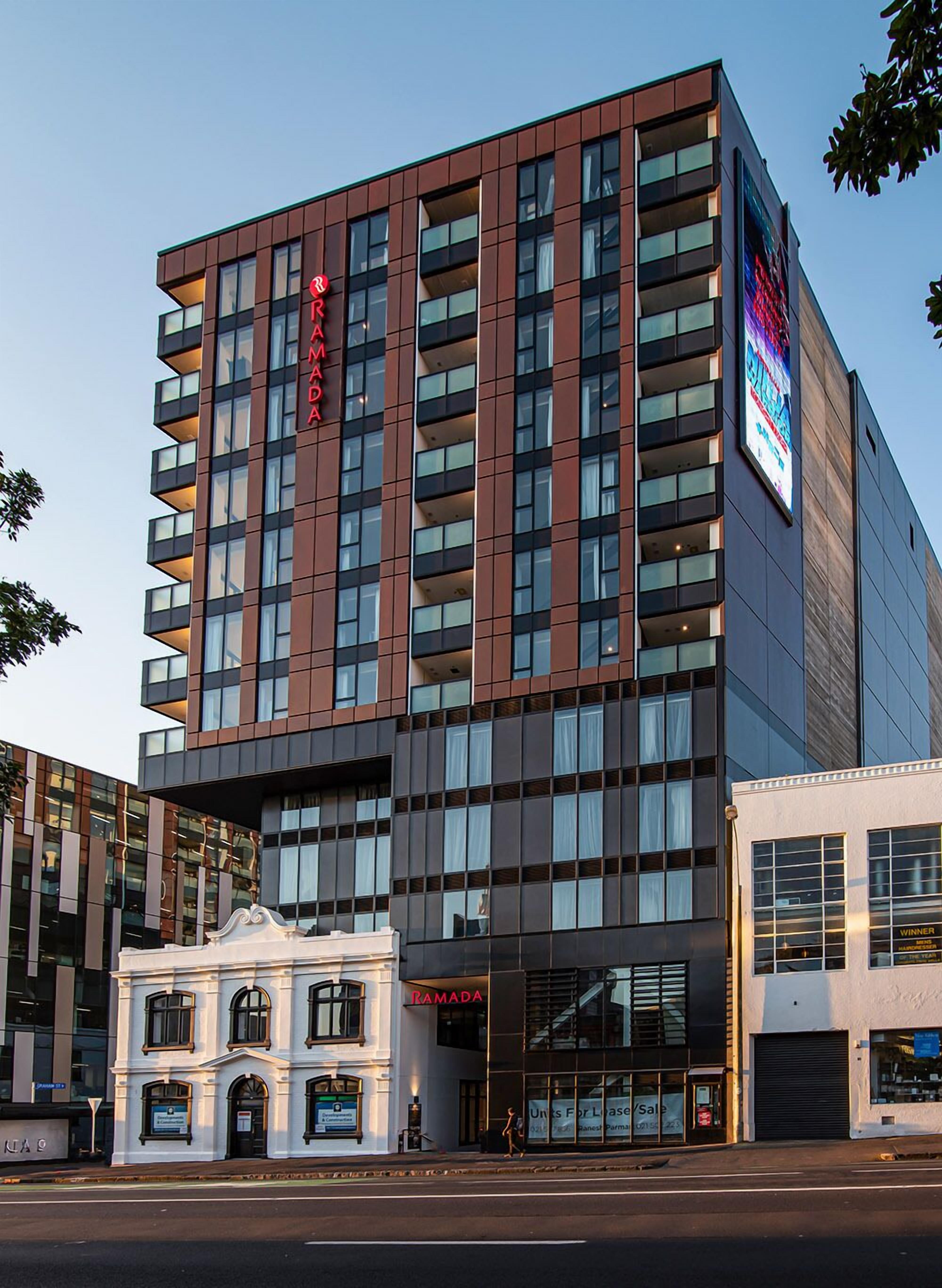 Ett modernt hotell med flera våningar, utmärkt av en tydlig skylt med texten ”RAMADA”. Fasadens design kombinerar glas och mörka paneler, och på markplan finns ett kommersiellt utrymme med en skylt som visar texten ”WINNER”.