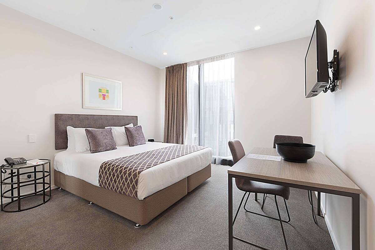Una habitación de hotel con cama, mesita de noche, silla, escritorio y televisión montada en la pared.