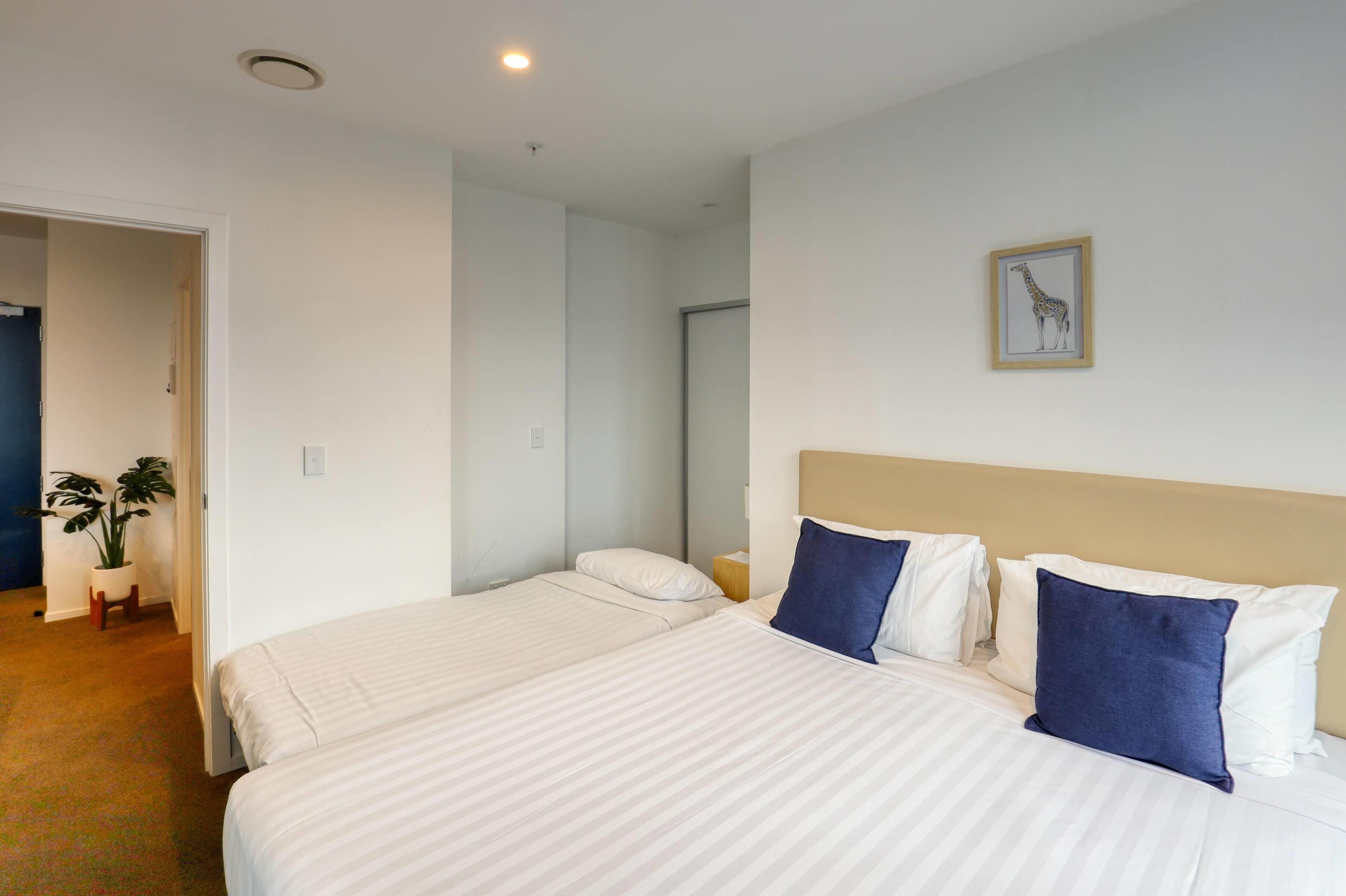 A hotel room with two beds, a large window, a plant, and a giraffe picture on the wall.