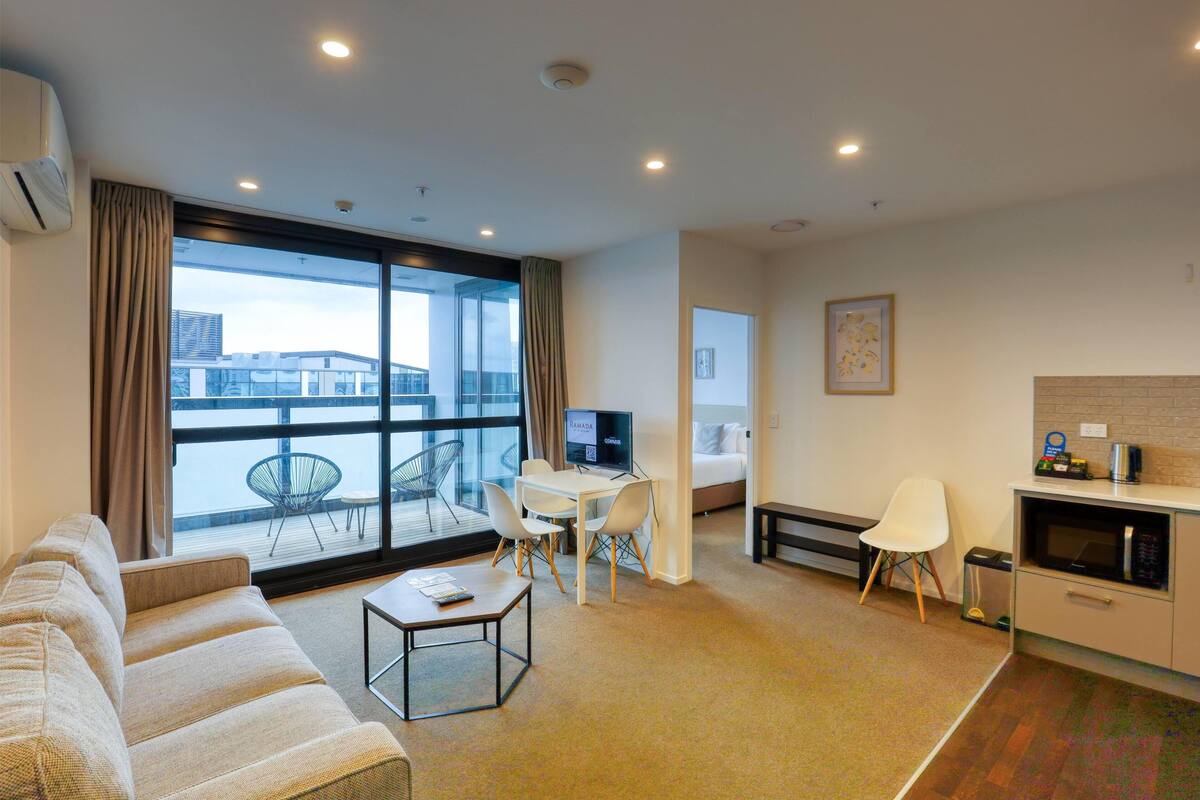 Una habitación de hotel moderna con un ventanal grande, un sofá, un comedor, un televisor y una kitchenette.