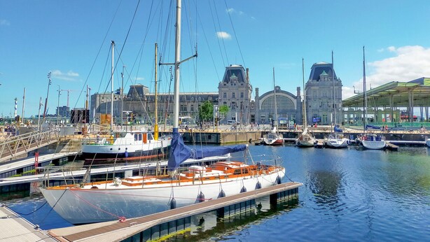 A marina with multiple boats docked, a large building with a distinctive arched entrance in the background, and a pedestrian bridge over the water. - Residentie Zeezicht, Ref 104 (Oostende)