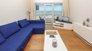 A modern living room with a blue sofa, a flat-screen TV, and a balcony with a view of the beach. - Residentie Zeezicht, Ref 104 (Oostende)