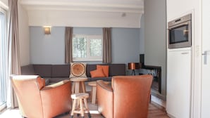 Un salon moderne avec un canapé gris, des fauteuils orange, une table basse ronde en bois et une grande fenêtre avec des rideaux.