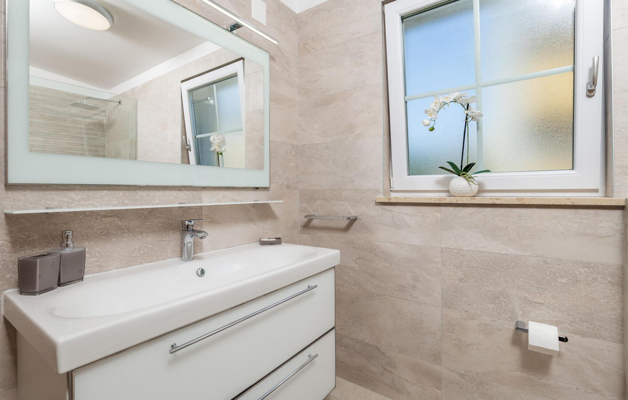 A modern bathroom with a white vanity, a large mirror, a window with a plant, and a towel dispenser.