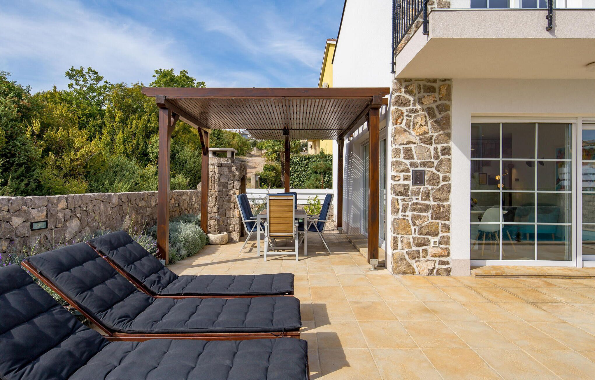 A patio with two lounge chairs, a pergola, and a stone wall.