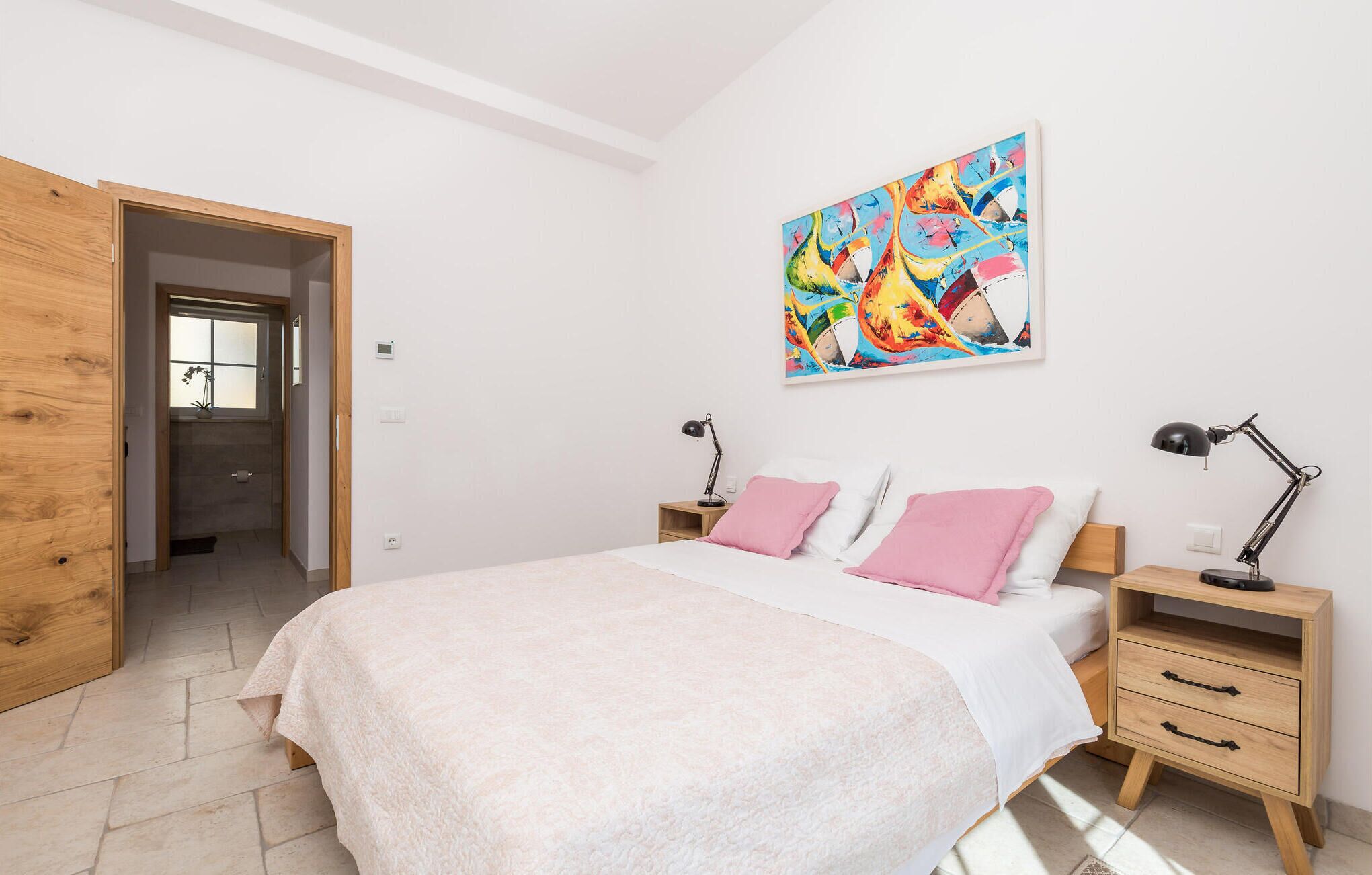 A bedroom with a bed, two bedside tables, a nightstand, a wooden wardrobe, a colorful abstract painting, and a potted plant.