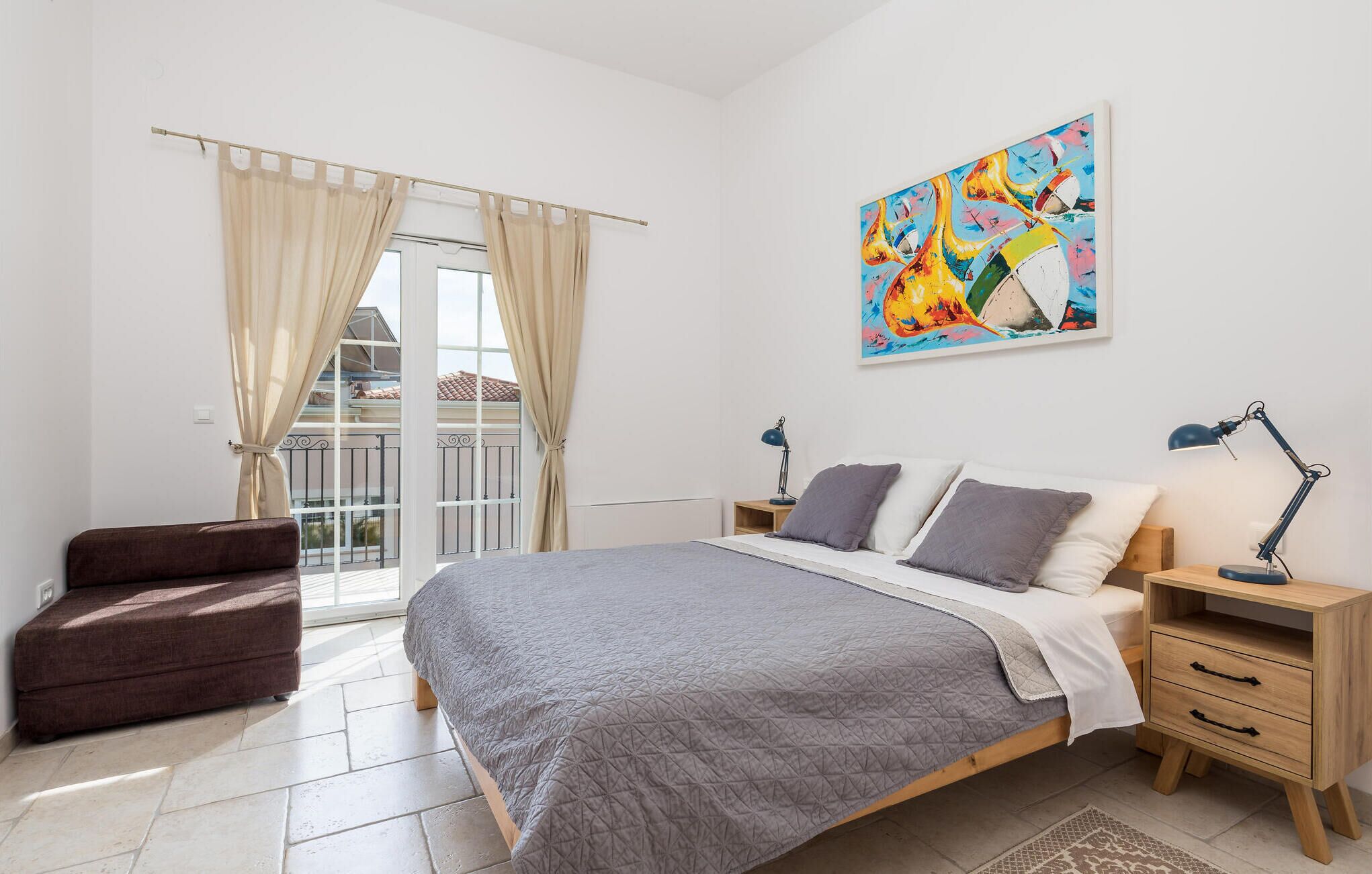 A bedroom with a bed, bedside tables, a nightstand, a chair, a window with curtains, and a colorful painting on the wall.