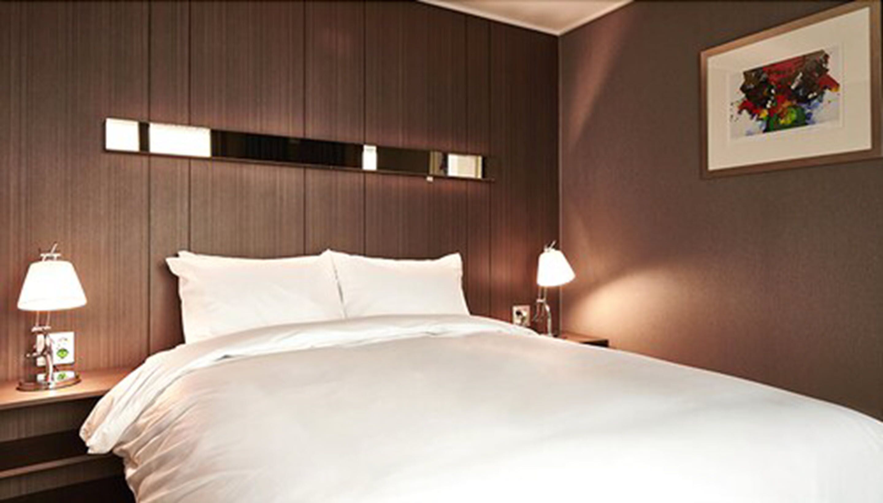 Habitación de hotel con una cama grande, dos mesitas de noche con lámparas, una obra de arte enmarcada en la pared y un diseño de paneles de madera en la pared.