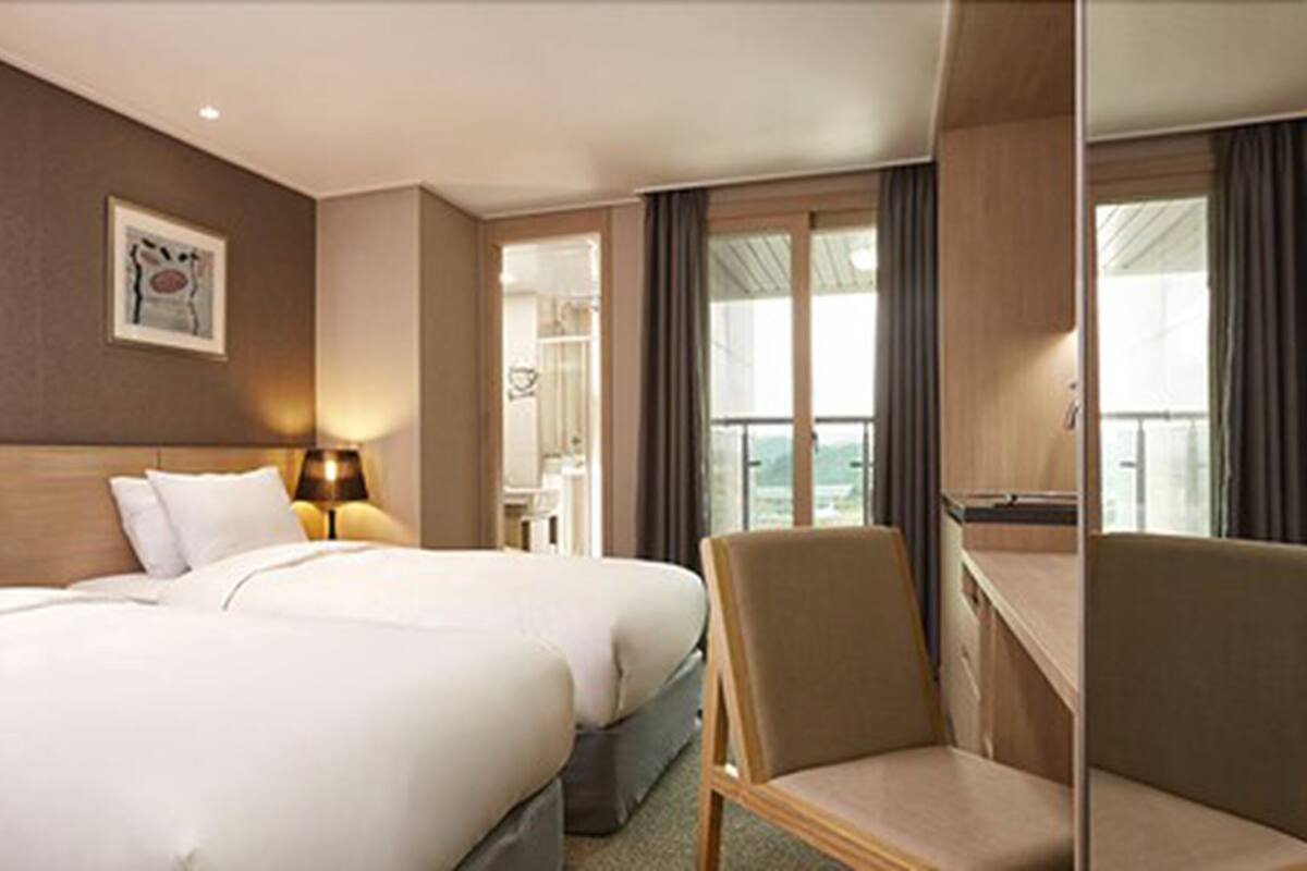 A hotel room with two beds, a chair, a desk, a lamp, and a window with curtains.