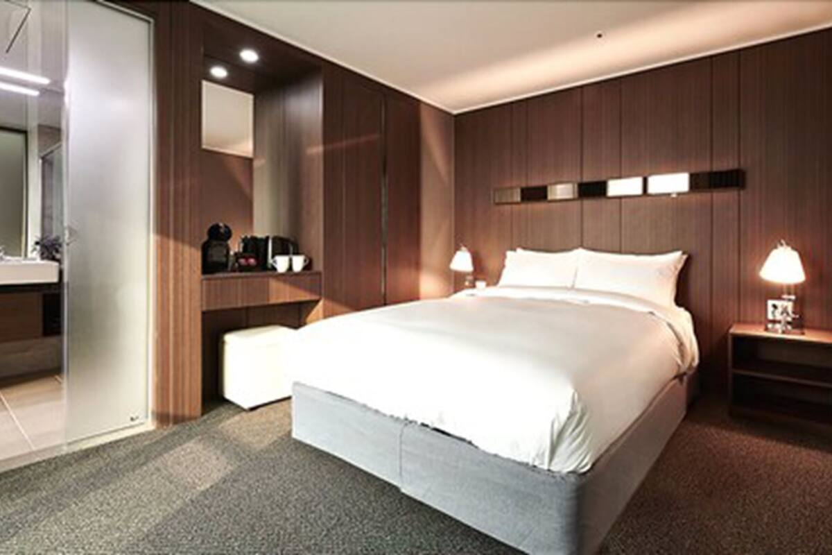 A modern hotel room with a large bed, bedside lamps, a small bedside table, a mirror, and a built-in shelf with toiletries.