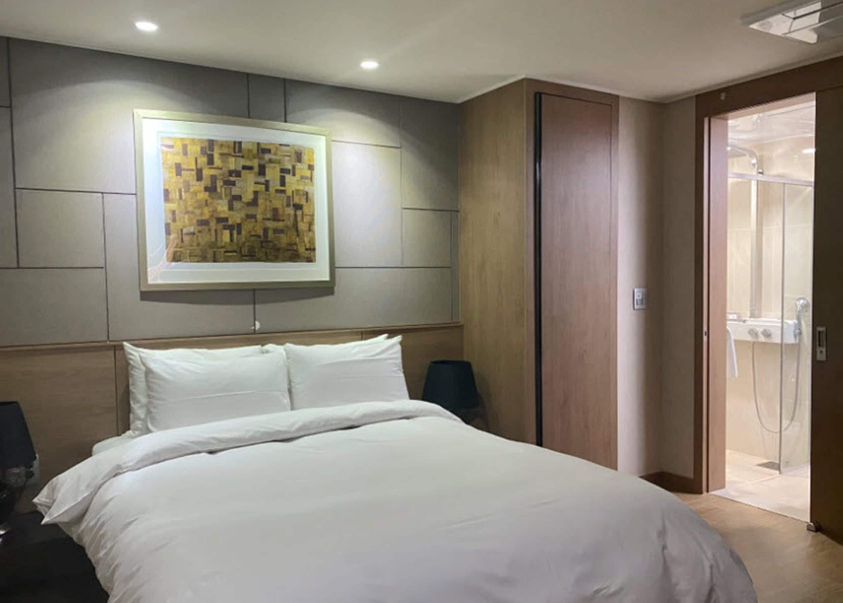 A hotel room with a large bed, a wooden headboard, a framed artwork on the wall, and a sliding door leading to a shower area.
