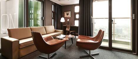 A modern hotel room with a sofa, two armchairs, a coffee table, and large windows offering a city view.