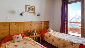 A room with two beds, a wooden headboard, a small table with a teddy bear and a lamp, a framed picture on the wall, and a window with a view of snow-capped mountains.