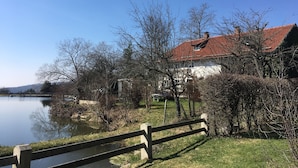 A riverside view with a wooden bridge, a white building with a red roof, and leafless trees.