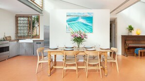 Un comedor con una mesa de madera dispuesta para seis comensales, un cuadro de una escena costera y una planta en maceta sobre el piano.