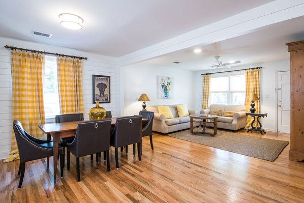 A living room with a dining area, wooden flooring, and yellow curtains. - Shem Creek Corner, one block from Historic Shem Creek waterway, beaches close (Mt Pleasant)