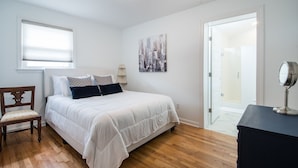 Een slaapkamer met een bed, een stoel, een nachtkastje, een spiegel, een schilderij en een deur naar een andere kamer.