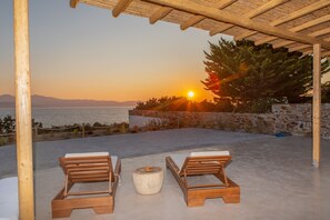 Eine Holz-Pergola mit zwei Liegestühlen und einem runden Tisch, mit Blick auf einen malerischen Sonnenuntergang über dem Meer.