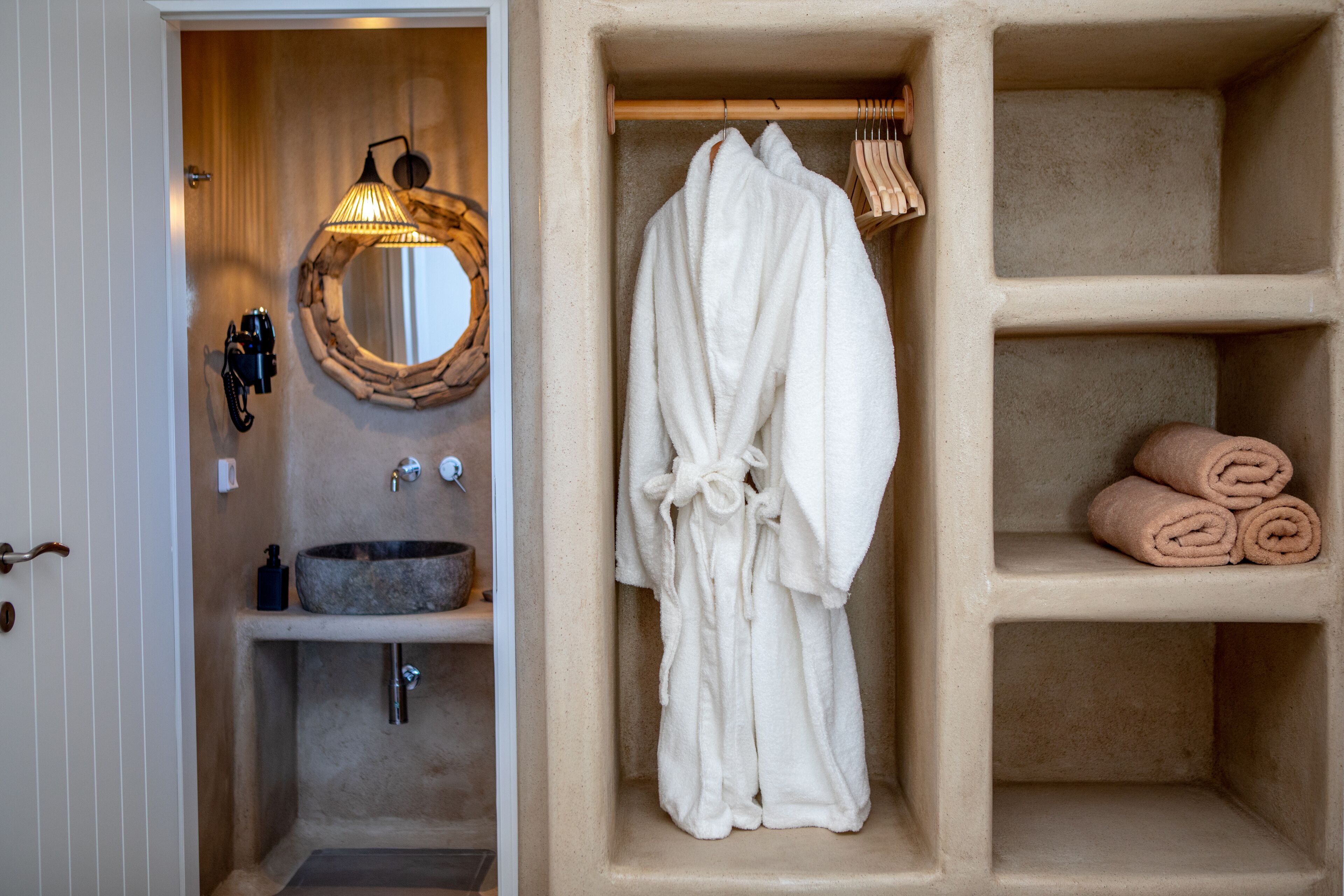 A bathroom with a stone sink, a wooden-framed mirror, a robe hanging on a rack, and shelves with rolled towels.