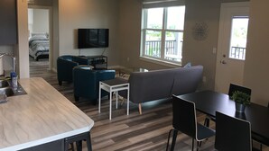 A modern kitchen with a central island, a dining area with a table and chairs, a living room with a sofa and a TV, and a balcony with a railing. - 2 bedroom + 1 bath apartment with amazing views! (Missoula)