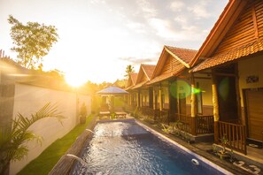 A swimming pool with lounge chairs and umbrellas, surrounded by wooden buildings with tiled roofs. - Myart Homestay (Penida Island)