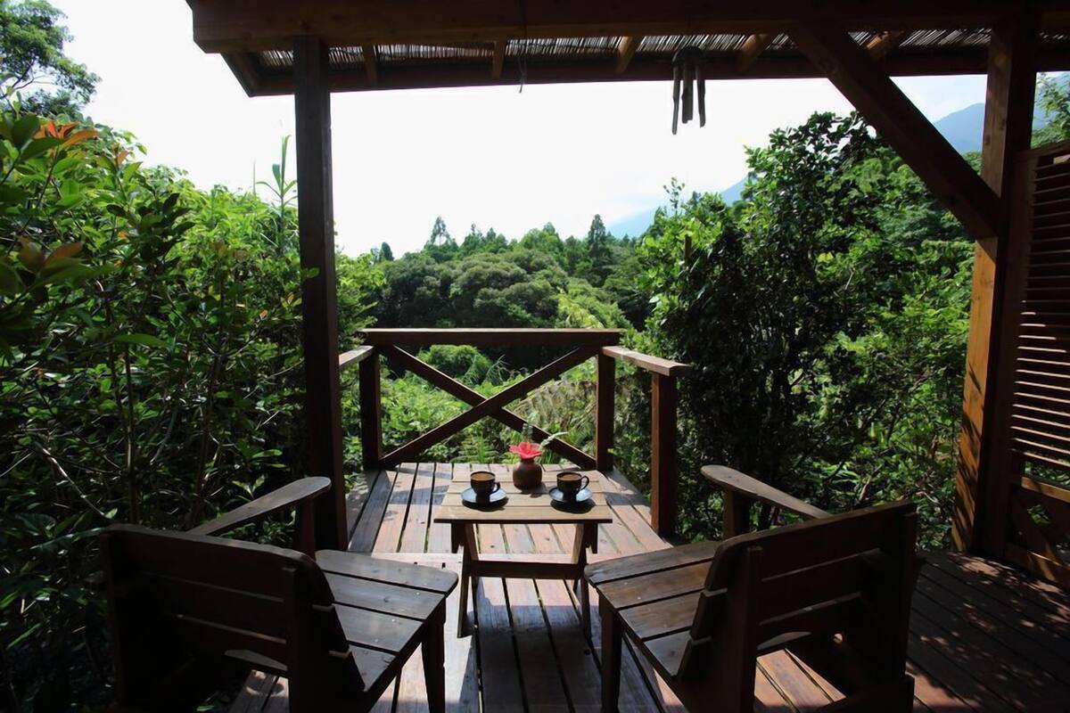 A wooden deck with a table and chairs, surrounded by lush greenery and a mountainous backdrop.