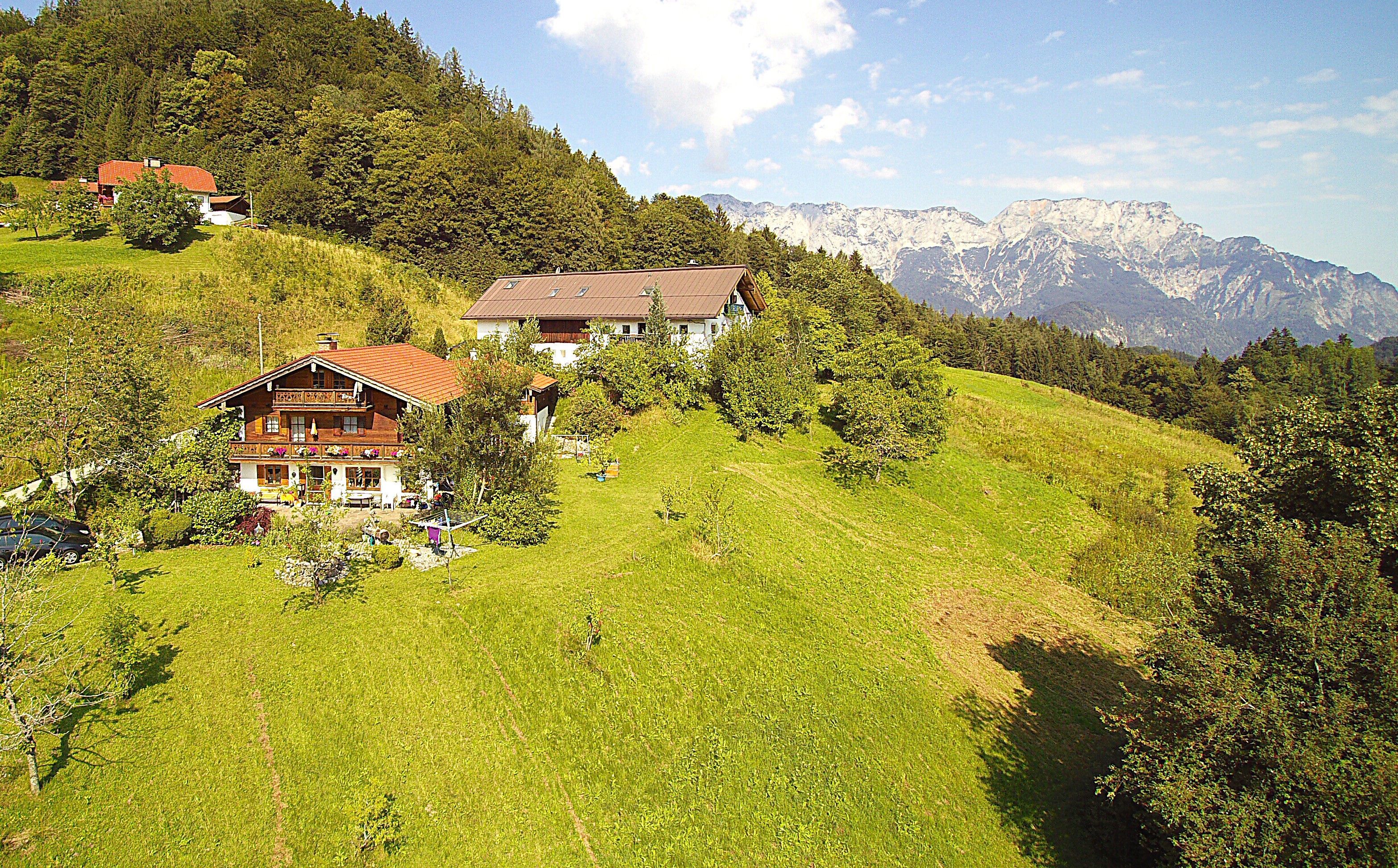 A hillside property with multiple buildings, including a chalet and a larger house with a balcony, surrounded by lush greenery and mountains in the background.
