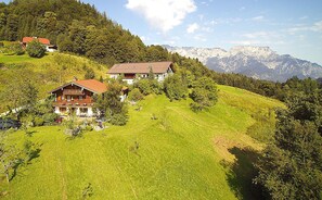 Een perceel op een heuvel met meerdere gebouwen, waaronder een chalet en een ruimer huis met een balkon, omgeven door weelderige begroeiing en bergen op de achtergrond.