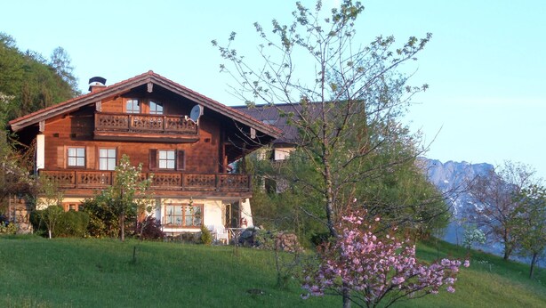 A wooden chalet with a balcony, surrounded by a grassy landscape and a flowering tree. - Vacation apartment Untersberg 38sqm (Berchtesgaden)