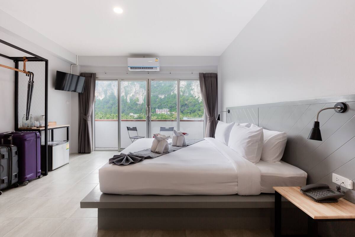 A modern hotel room with a large bed, a desk with a phone, a TV, and a balcony with a view of greenery.