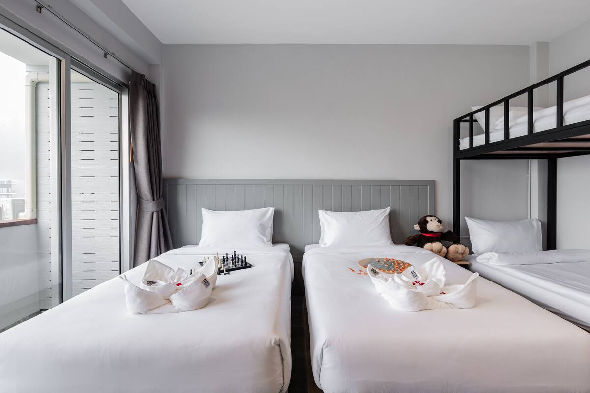 A hotel room with two single beds, a bunk bed, a window with blinds, and a stuffed monkey toy on one bed.