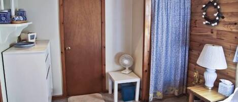 A compact room with a wooden door, a small table with a lamp, a bed, and a fan on a small table.