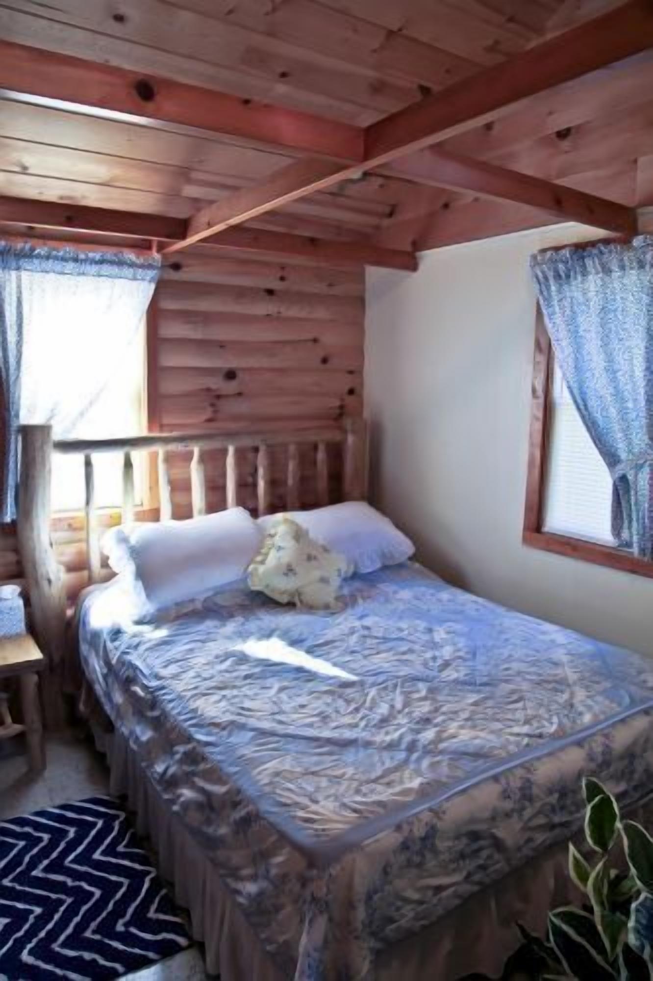 A wooden cabin room with a bed, a window with curtains, a wooden desk, and a patterned rug.